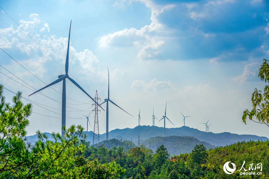 青山綠樹間，在藍天白云的映襯下，多座風力發電機與輸電塔矗立在山脊上。人民網 涂詩軒攝